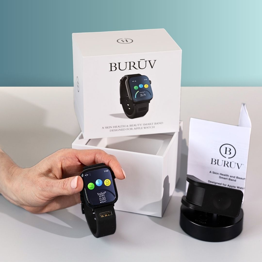 A hand holds a black BURŪV smart band for Apple Watch® in front of its white product box and charging dock on a light gray surface against a teal-blue background.
